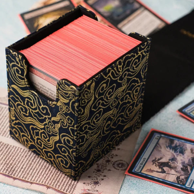 A Magic: The Gathering card deck box with a black and gold cloud-patterned fabric cover, filled with red cards, styled on an old book over a light blue textured surface.