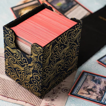 A Magic: The Gathering card deck box with a black and gold cloud-patterned fabric cover, filled with red cards, styled on an old book over a light blue textured surface.