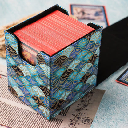 A Magic: The Gathering card deck box with a blue and teal wave pattern, filled with red cards, displayed on a light blue textured surface with cards and a book spread around it.
